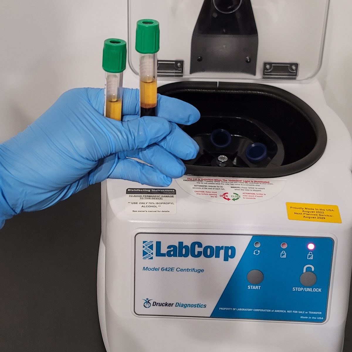 A blue-gloved hand holding two green-capped blood tubes, each with a separated golden layer on top, beside an open LabCorp Model 642E benchtop centrifuge