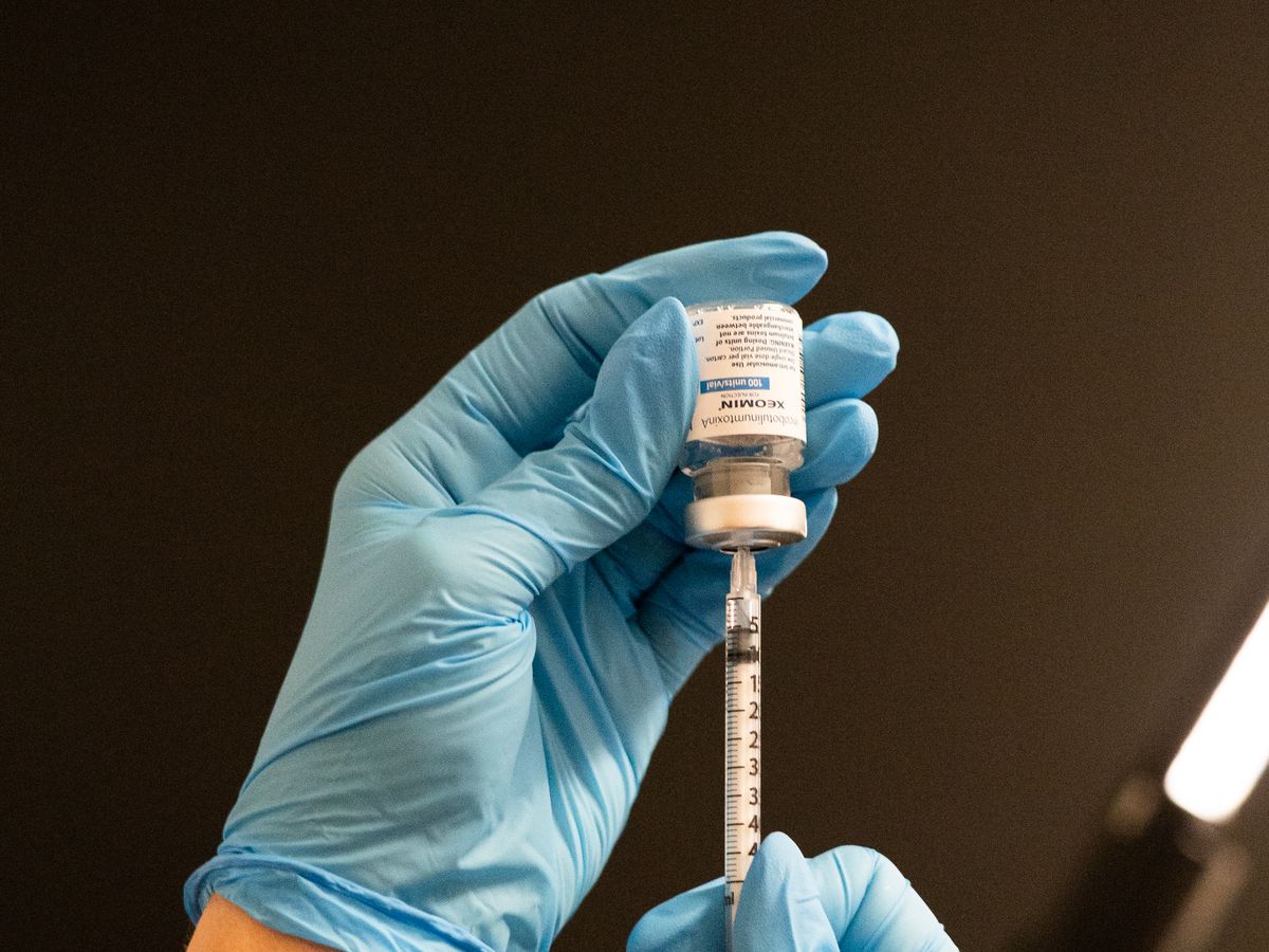 Two hands in blue nitrile gloves drawing liquid from an upturned Xeomin vial into a graduated syringe against a dark background