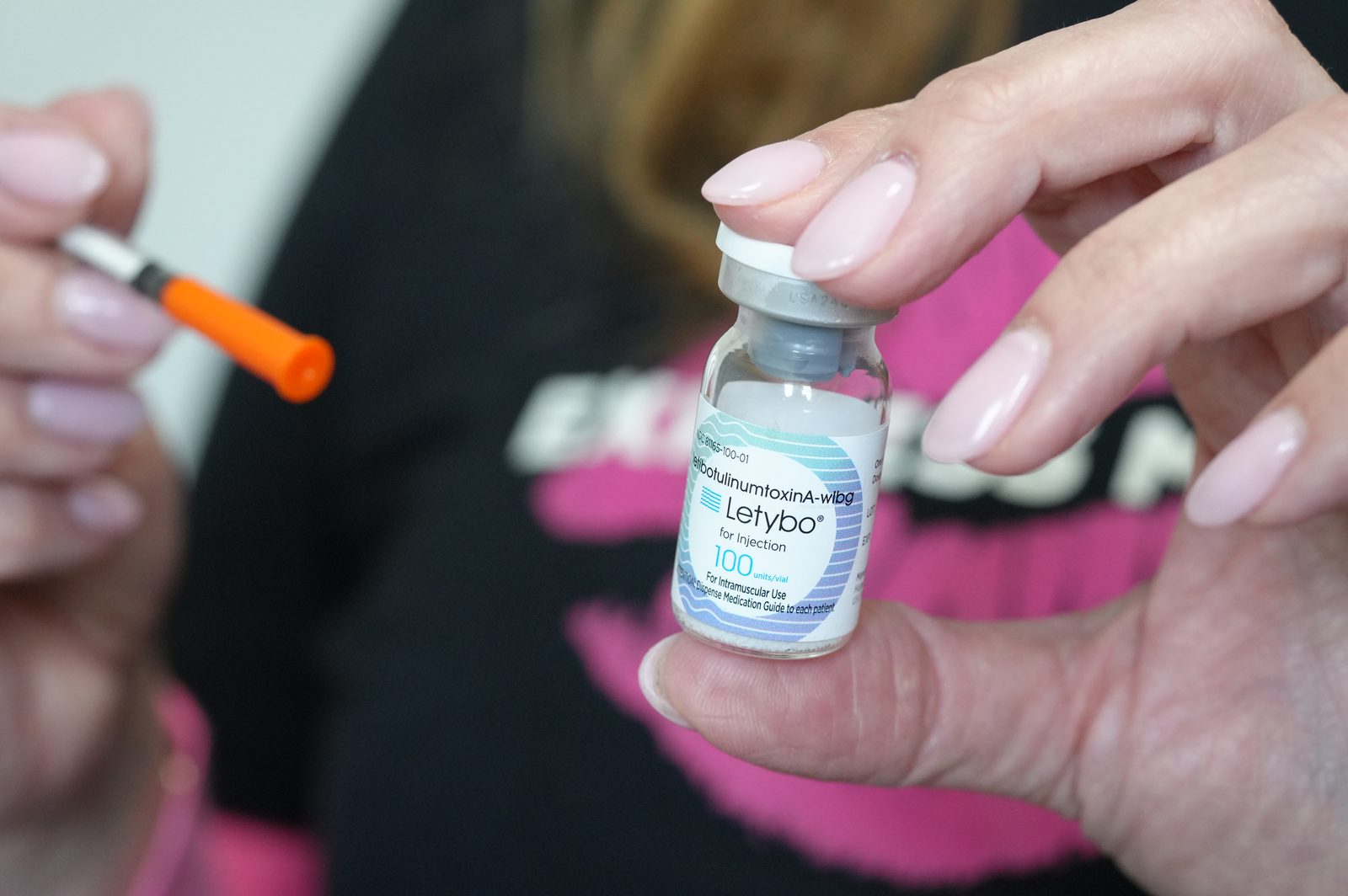 A hand with manicured nails holding a Letybo 100 Units vial up to the camera, an orange-capped syringe in the other hand and a black shirt blurred behind