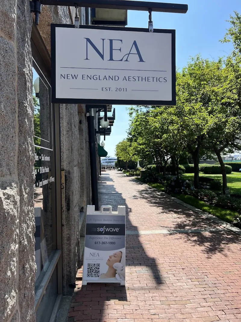 Hanging New England Aesthetics sign over a brick walkway beside a granite building