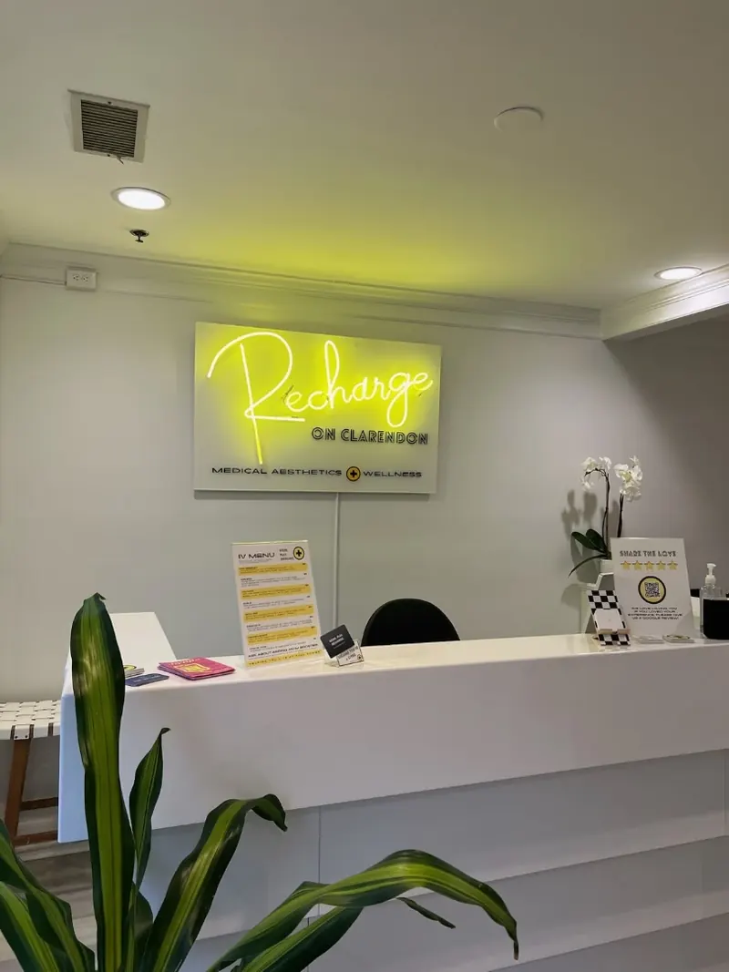 White reception desk beneath a yellow neon Recharge on Clarendon wall sign