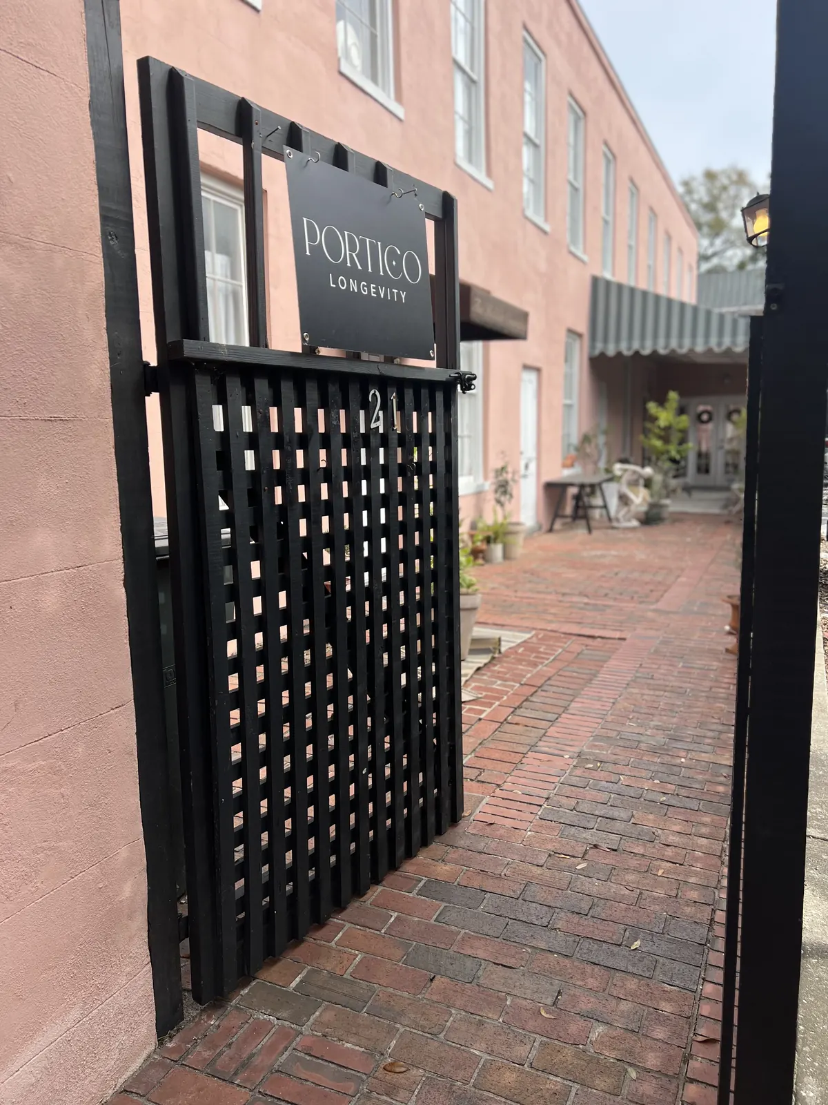 Black lattice gate with a Portico Longevity sign and number 21 over a brick path