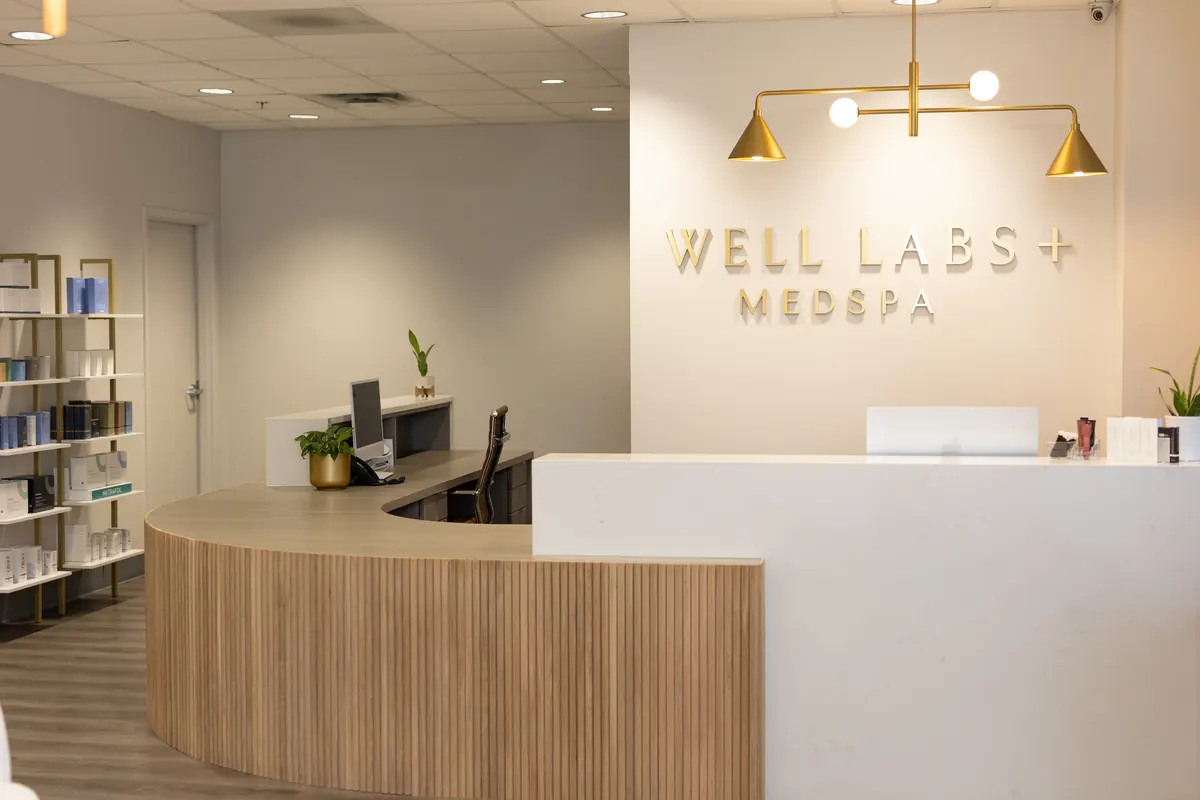 Curved wood and white reception desk below gold Well Labs+ Medspa wall lettering