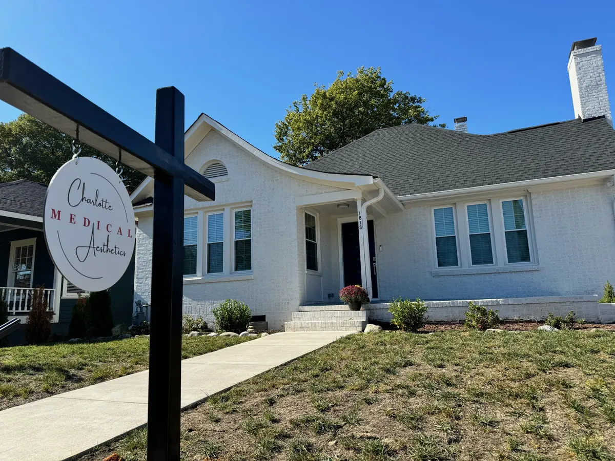 White brick bungalow with a front door and a hanging Charlotte Medical Aesthetics sign on a post