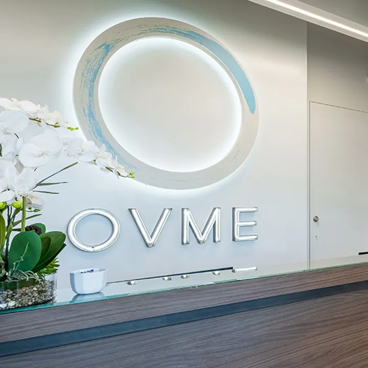 Backlit OVME ring logo and silver lettering behind a glass-topped reception counter with orchids