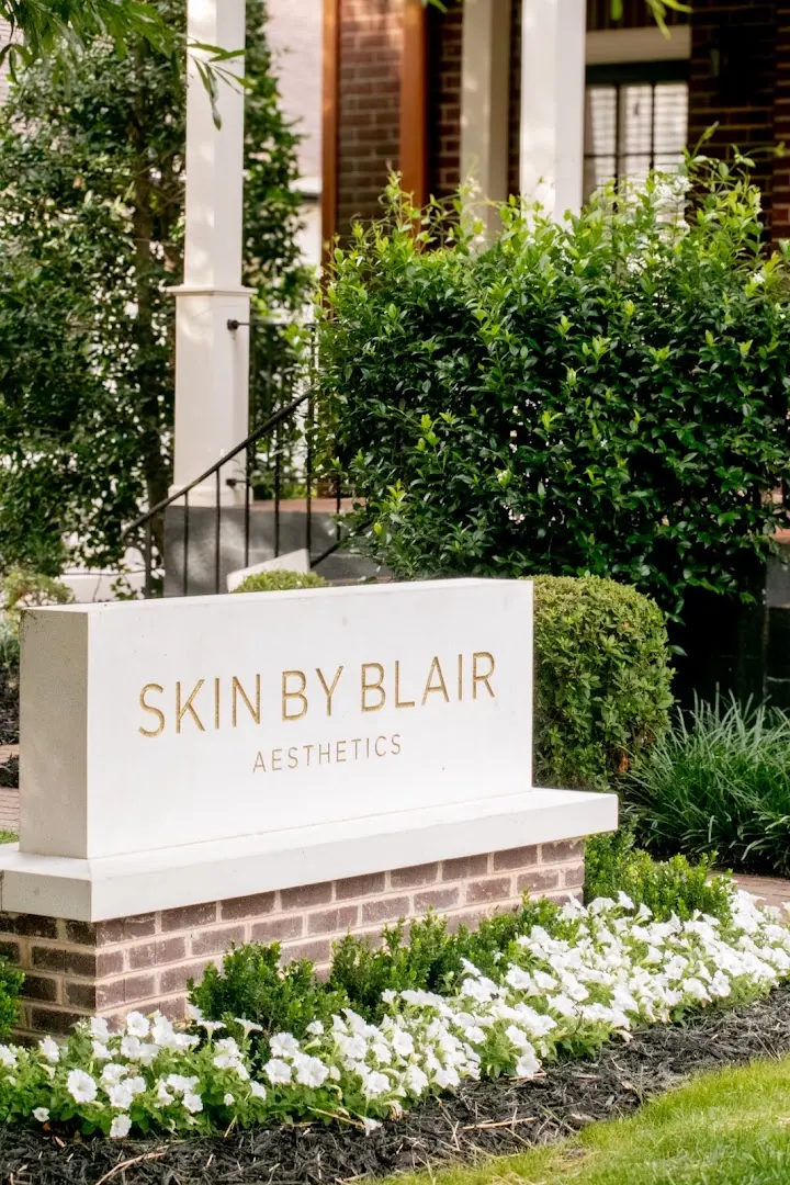 Stone monument sign reading Skin By Blair Aesthetics beside white flowers and a hedge