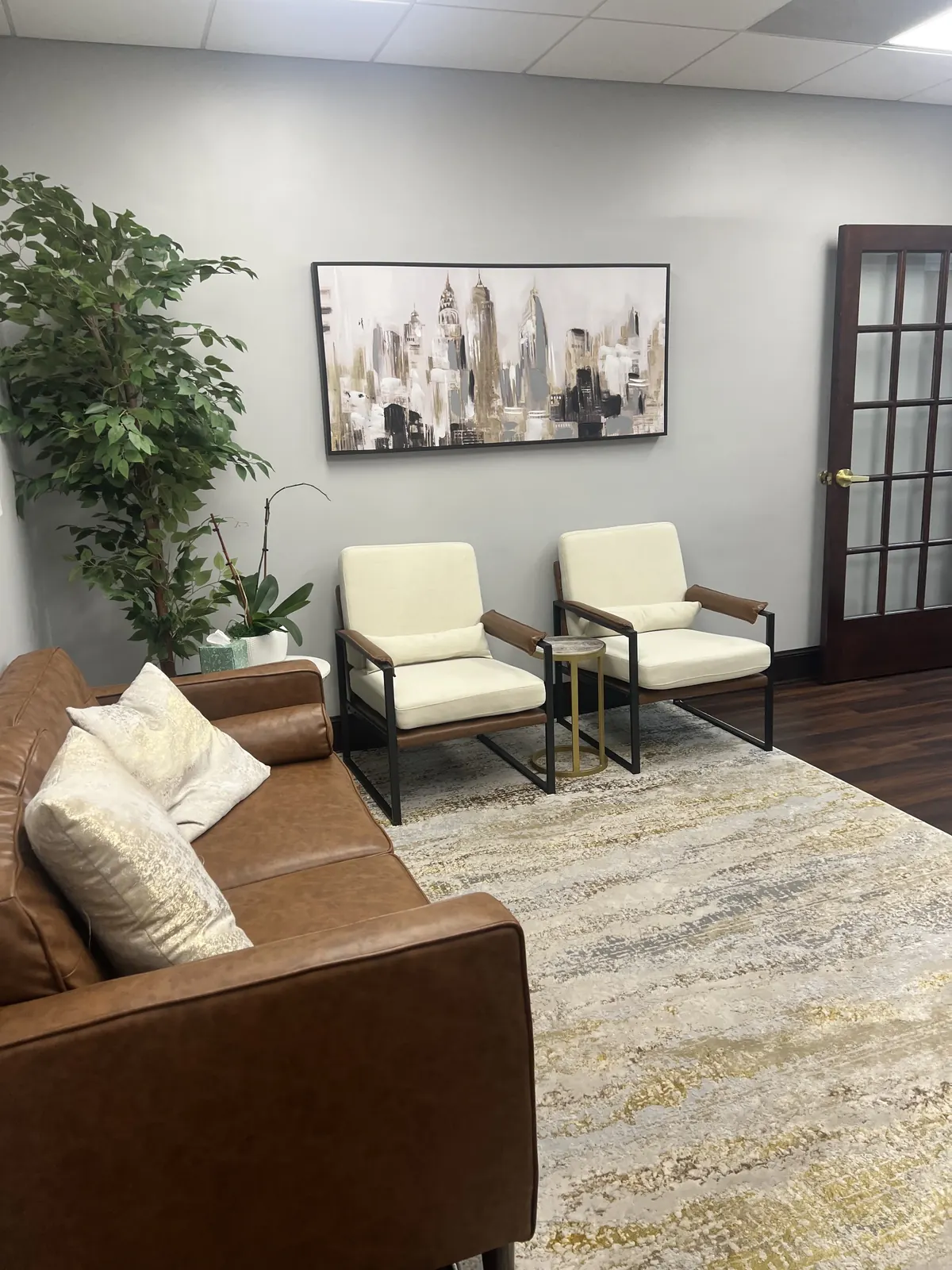 Waiting area with a brown leather sofa, two cream armchairs and framed skyline art