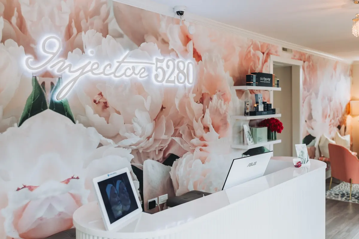 Reception desk under a lit Injector 5280 neon sign on a pink peony mural