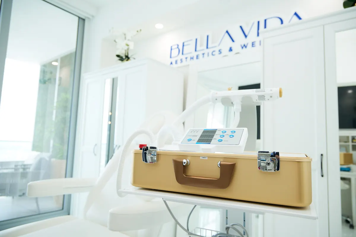 Gold-cased device on a cart in a white treatment room beneath a Bella Vida sign