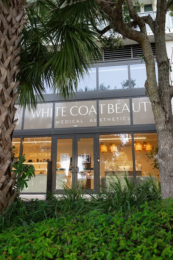 Glass storefront with White Coat Beauty Medical Aesthetics lettering behind palms and shrubs