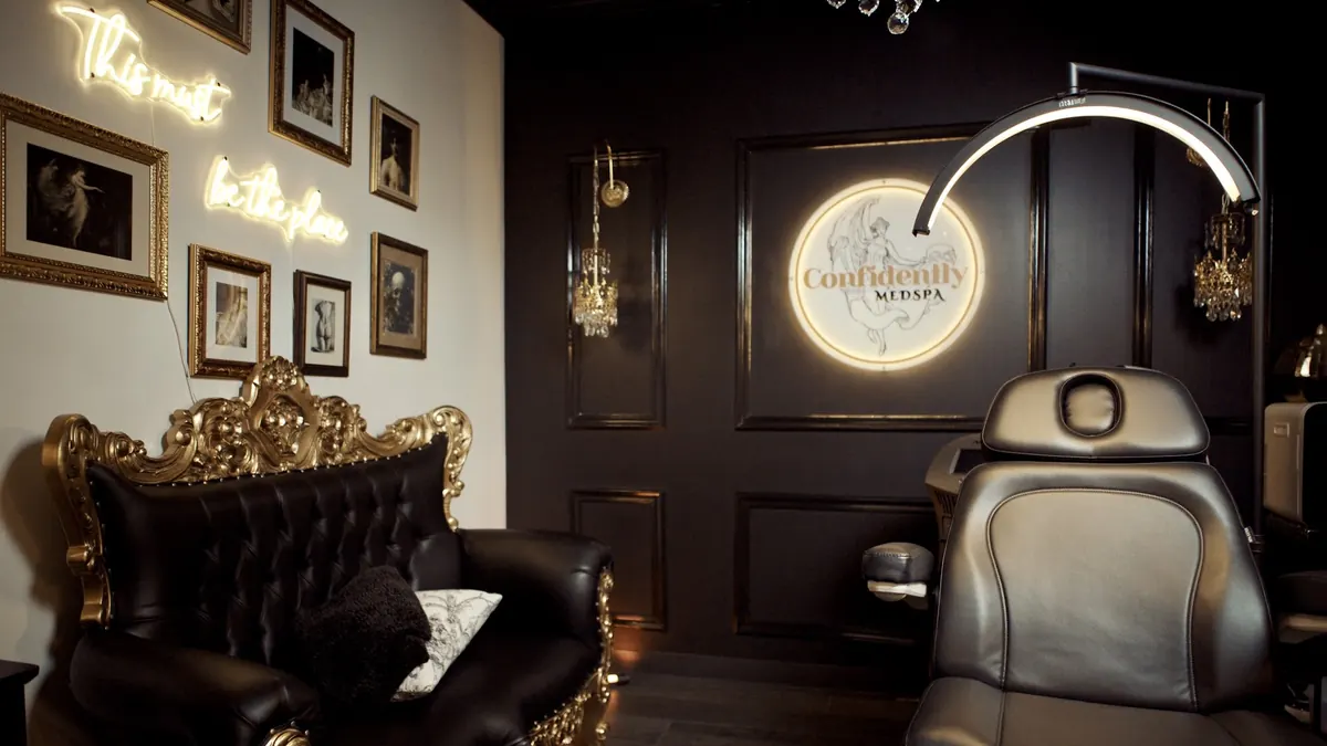 Dark paneled treatment room with a lit Confidently Medspa logo, treatment chair and gold-framed sofa