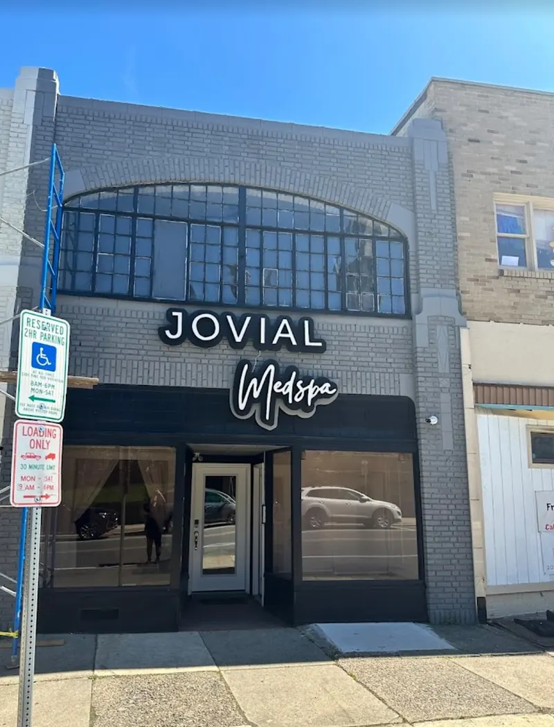 Gray brick storefront with a glass door under an illuminated Jovial Medspa sign and arched upper windows