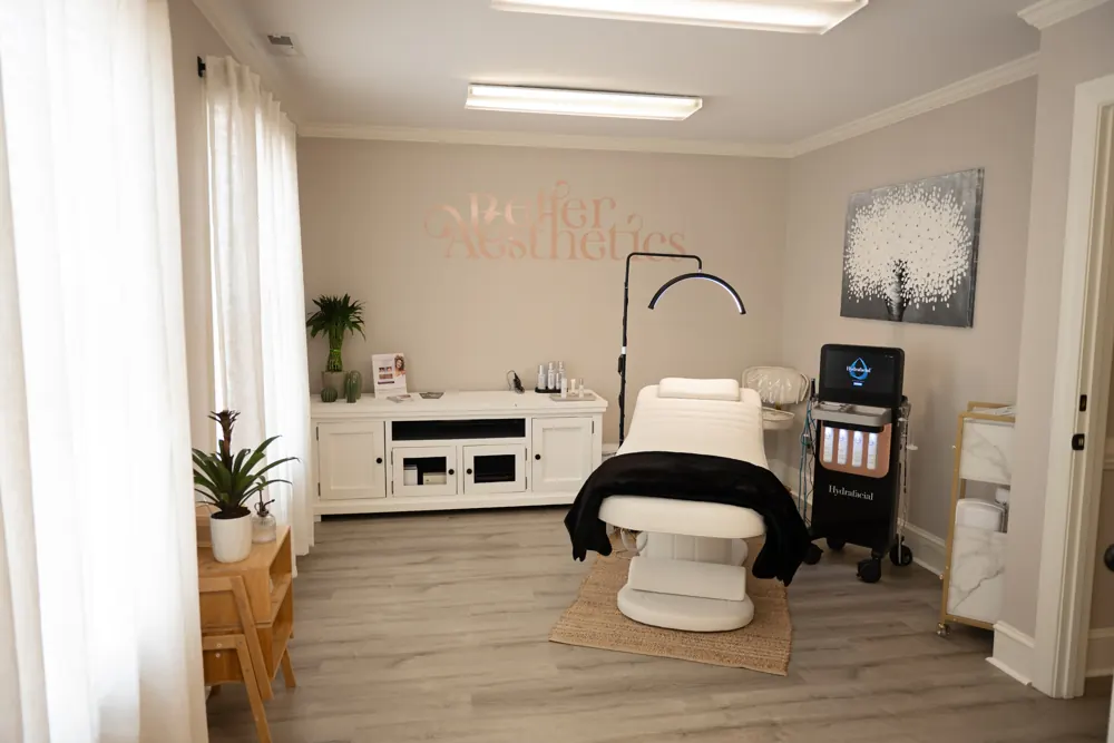 Treatment room with a padded treatment chair, device cart and rose-gold Better Aesthetics wall lettering