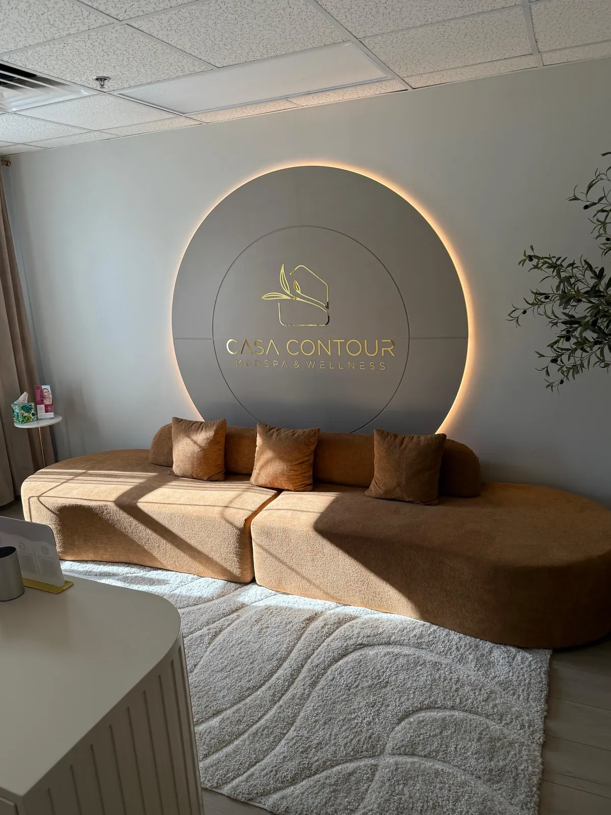 Curved tan sofa and rug beneath a backlit circular wall logo reading Casa Contour