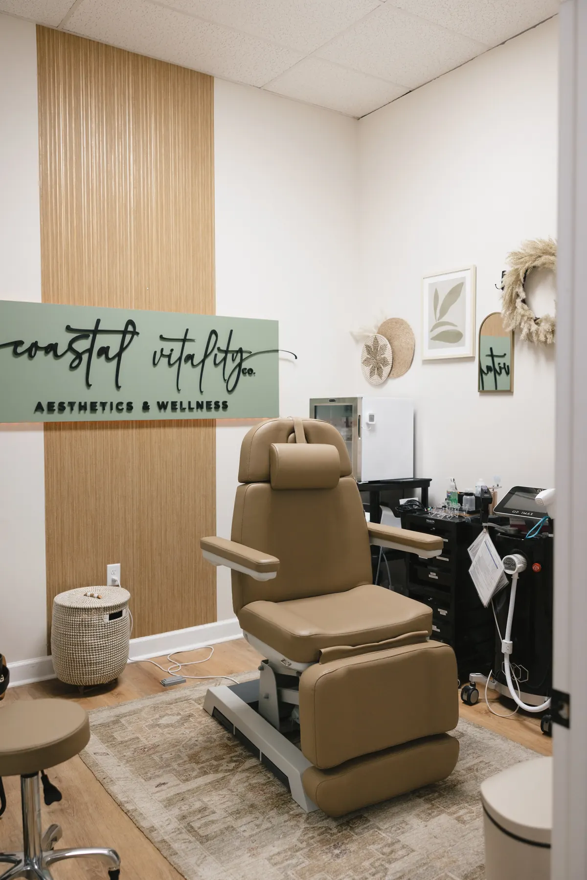 Tan treatment chair, equipment cart and wood slat wall beneath a green coastal vitality co. sign