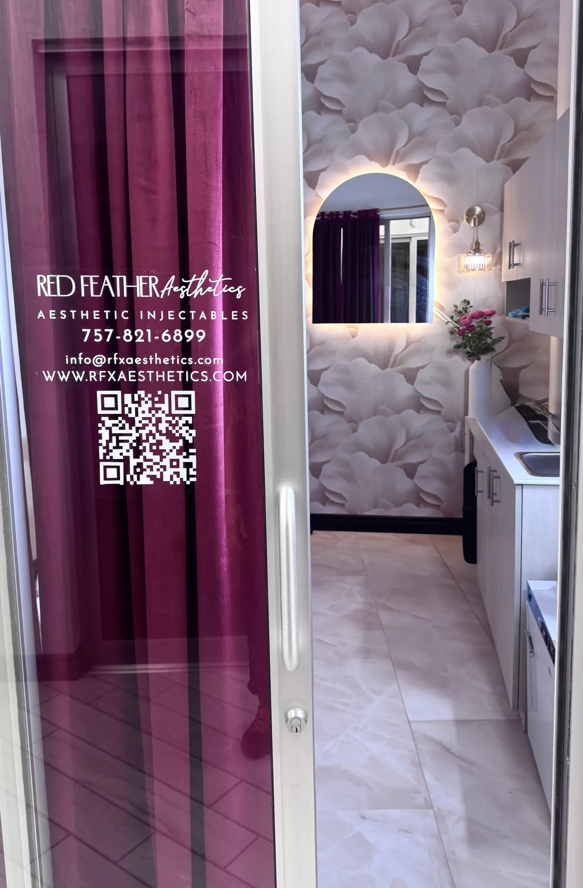 Glass door with a Red Feather Aesthetics decal, with a floral-wallpapered treatment room behind